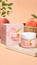 A jar of Iconsign Cleansing Balm in peach flavor sits next to its pink packaging box on a light stone slab, with peaches and green leaves blurred in the background against a peach-colored backdrop.