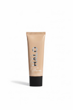 A tube of HALO Healthy Glow All-In-One Tinted Moisturizer SPF 20, in a beige shade, is displayed on a glass counter inside a retail store. A hand is holding a white tablet in the background, and various store fixtures and other products are visible.