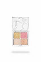 A hand holding an open Dior 4-pan eyeshadow palette. The clear plastic case features a large silver-tone Dior logo on the inside of the lid, and the four eyeshadow pans are arranged in a grid with gold, pink, champagne, and peach tones, all embossed with the Dior Oblique pattern.
