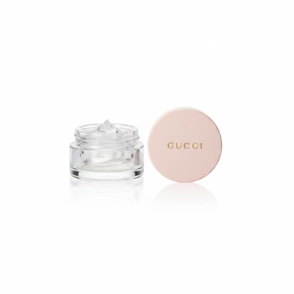 A Gucci round cosmetic compact with a pale pink lid and a clear jar with a silver-toned product, placed on a glass tabletop next to mannequin heads and a red patterned box.