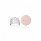 A Gucci round cosmetic compact with a pale pink lid and a clear jar with a silver-toned product, placed on a glass tabletop next to mannequin heads and a red patterned box.