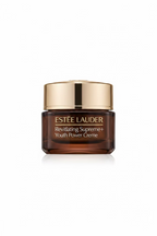A hand holding an Estée Lauder Revitalizing Super Cream jar, showing the gold and clear packaging against a background of clothing and storage items.