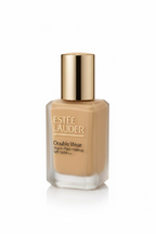 Estée Lauder Double Wear Stay-in-Place Makeup SPF 10/PA++ foundation bottle with a metallic silver cap, placed on a reflective glass surface with a laptop and red folder in the background.
