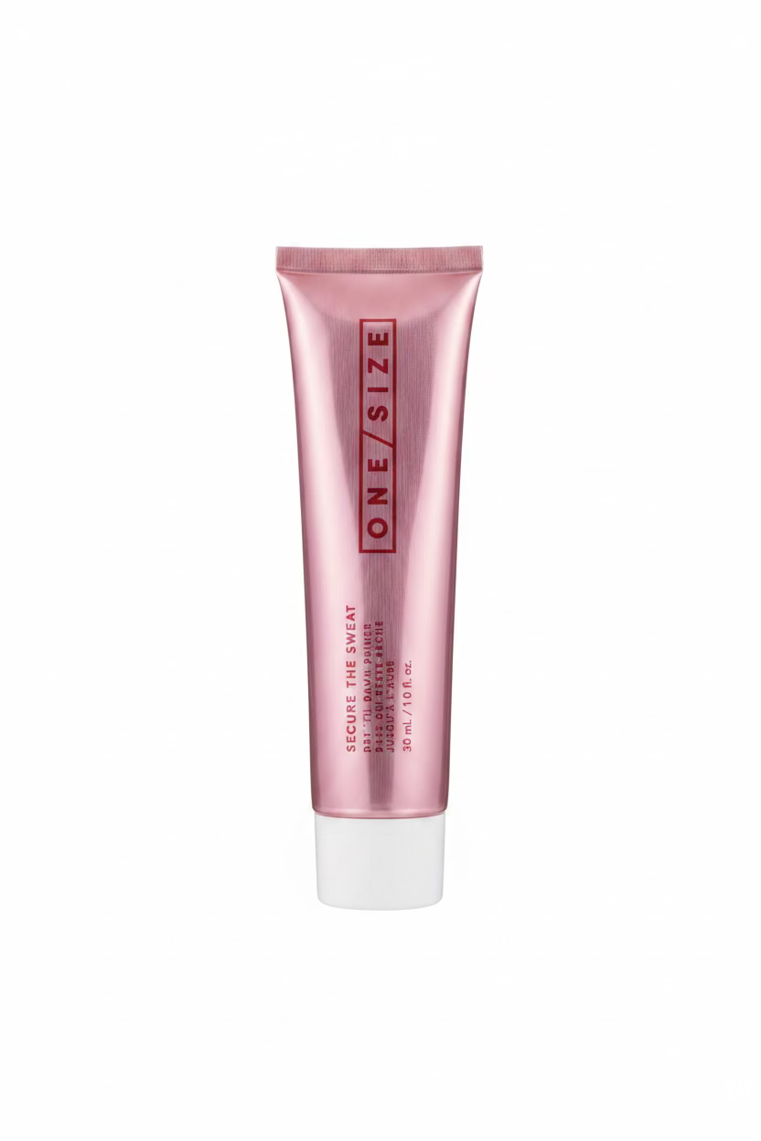 A pink squeeze tube of One Size Secure the Sweat hair product standing upright in front of a yellow plastic basket on a glass table, with shelves of boxes in the background.