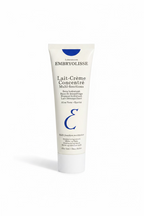 A hand holding a white tube of Embryolisse Lait-Crème Concentré Multi-fonctions moisturizer cream, with a person's legs and jeans visible in the background.
