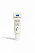 A hand holding a white squeeze tube of Laboratoires Embryolisse Lait-Crème Concentré Multi-fonctions moisturizer cream, with a person's jeans and shoes visible in the background.