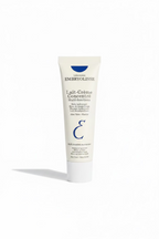 A hand holding a white squeeze tube of Laboratoires Embryolisse Lait-Crème Concentré Multi-fonctions moisturizer cream, with a person's jeans and shoes visible in the background.