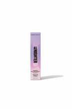 A box of Urban Decay All Nighter Maximum Cover Spray in the Matte finish, standing on a glass display counter inside a retail store. The pink and purple packaging is clearly visible, with other products and a laptop in the background.