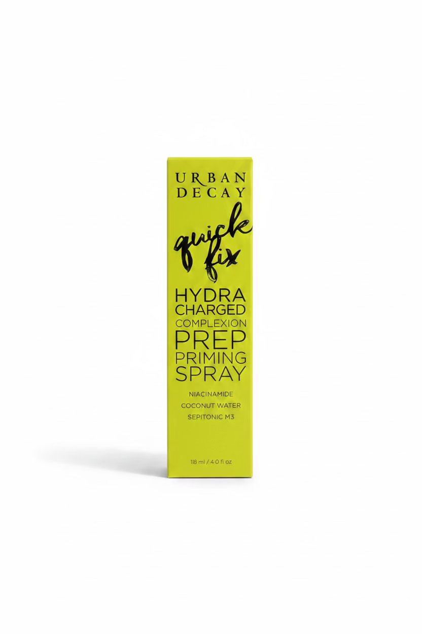 A yellow box of Urban Decay Quick Fix Hydra Charged Prep Priming Spray stands on a glass display counter in a retail store. Winston and Mynk boxes are visible in the background, along with a laptop and other cosmetics.