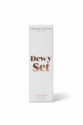 A white box of Anastasia Beverly Hills Dewy Set Setting Spray standing on a glass display counter in a retail store. The box features the brand name 'ANASTASIA BEVERLY HILLS' and the product name 'Dewy Set' in brown lettering.