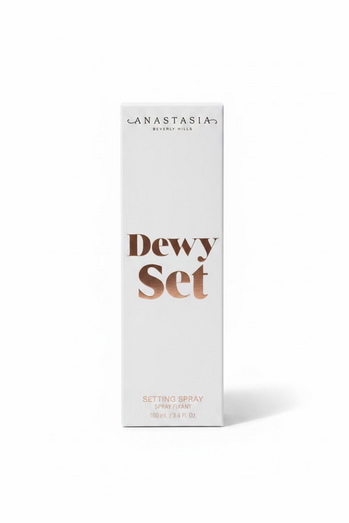A white box of Anastasia Beverly Hills Dewy Set Setting Spray standing on a glass display counter in a retail store. The box features the brand name 'ANASTASIA BEVERLY HILLS' and the product name 'Dewy Set' in brown lettering.