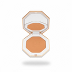 A hand holding an open octagonal makeup compact with a mirror on the lid and a warm tan pressed powder pan featuring an embossed 'FB' logo. The compact has a white and pale pink frame with gold-toned edges, set against a striped shirt and a cluttered indoor background.