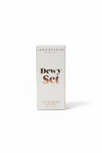 A white box of Anastasia Beverly Hills Dewy Set Setting Spray standing on a glass table, with a laptop and store items blurred in the background.