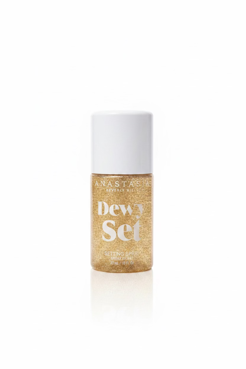 A bottle of Anastasia Beverly Hills Dewy Set Setting Spray with a white pump and a gold glitter bottle, sitting on a glass shelf in front of a laptop.