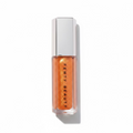 A hand holding a tube of Fenty Beauty liquid lipstick in a vibrant orange shade, with a silver cap, set against a background of denim jeans and a wooden surface.