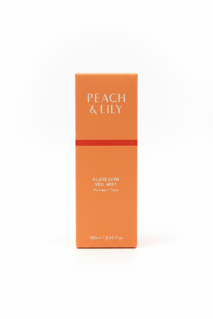 A peach-colored box of Peach & Lily Glass Skin Veil Mist stands on a glass display shelf, surrounded by other cosmetic packaging in a store setting.