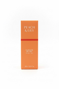 A peach-colored box of Peach & Lily Glass Skin Veil Mist stands on a glass display shelf, surrounded by other cosmetic packaging in a store setting.