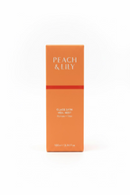 A peach-colored box of Peach & Lily Glass Skin Veil Mist stands on a glass display shelf, surrounded by other cosmetic packaging in a store setting.