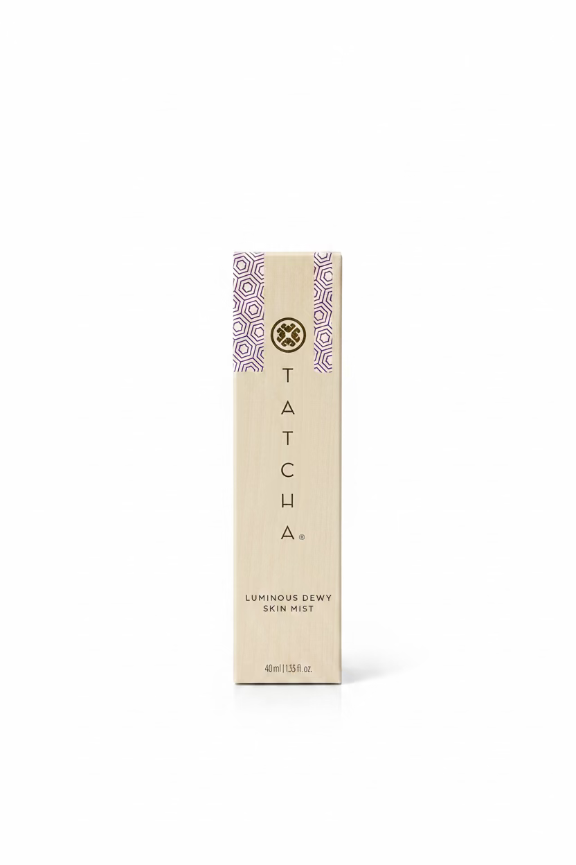A tall, narrow beige box for Tatcha Luminous Dewy Skin Mist stands on a glass display counter inside a retail store. The box features the Tatcha logo and name vertically printed in black, with a decorative blue and white patterned stripe on the right side.