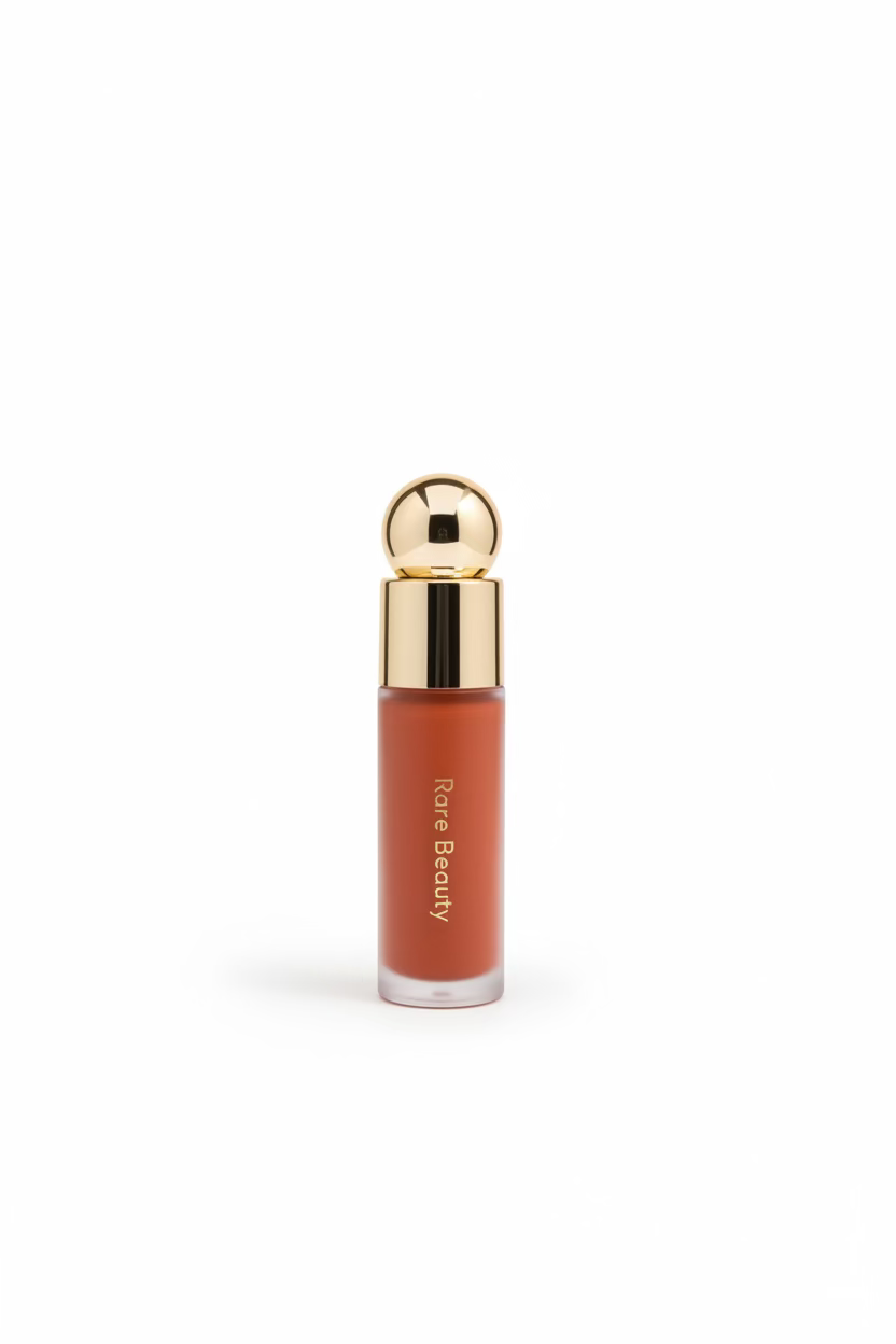 A Rare Beauty liquid blush in a clear bottle showing a warm peachy-orange shade, topped with a shiny gold cap and spherical ornament, set on a glass counter with a laptop and store items in the background.