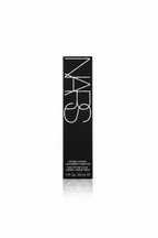 A hand holding a single NARS Natural Radiant Longwear Foundation tube in its original black packaging, with a beige shirt and a store shelf in the background.
