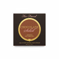 A box of Too Faced Chocolate Soleil Combed Matte Bronzer powder, placed on a glass table next to a laptop and a person's jeans.