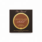 A box of Too Faced Chocolate Soleil Combed Matte Bronzer powder, placed on a glass table next to a laptop and a person's jeans.