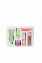 A boxed set of six cosmetic products arranged in a white tray, photographed on a glass display counter in a retail store. The set includes green, pink, and beige tubes and jars, with visible brand text including Glow Ron and Extrait de Beauté.