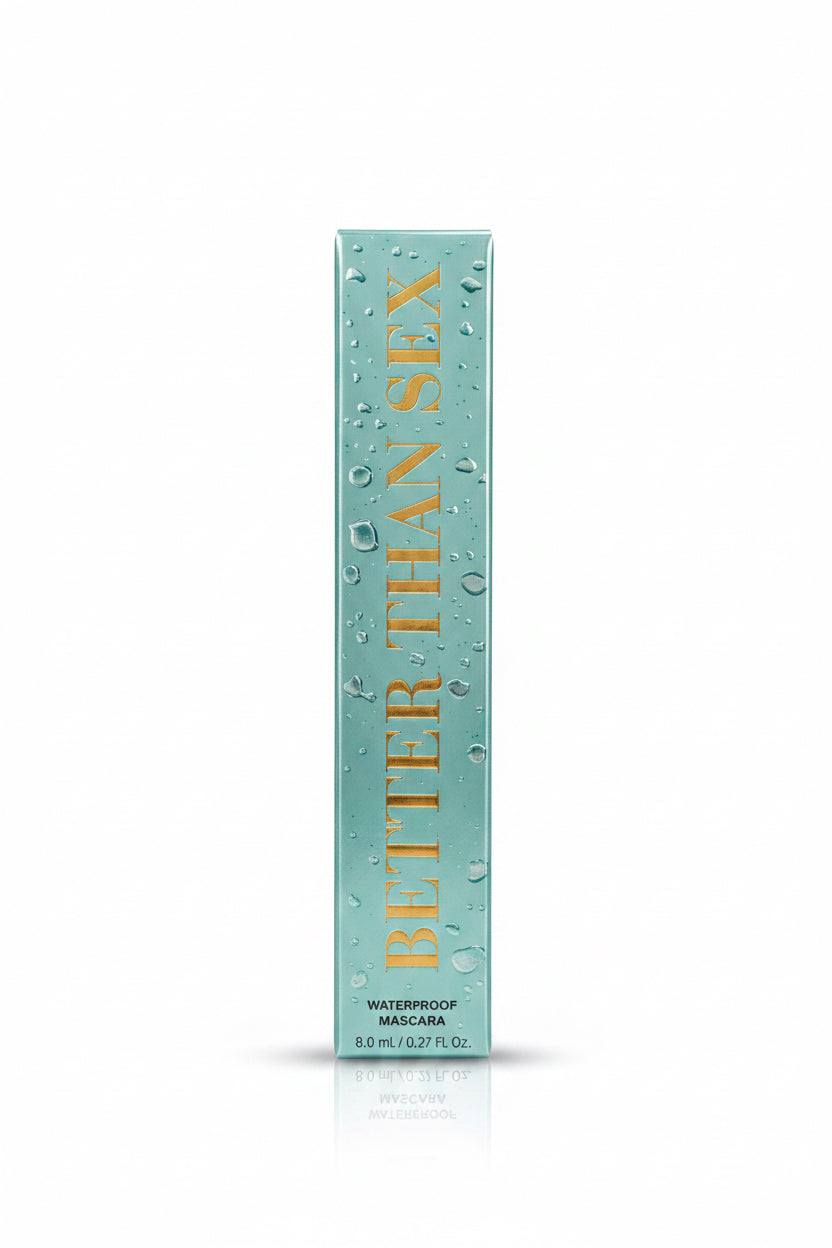 A boxed cosmetic product labeled 'Better Than Sex' stands upright on a reflective counter, with a gold promotional insert behind it. A person in a black shirt is visible in the background holding a phone, and a laptop is open on the counter.