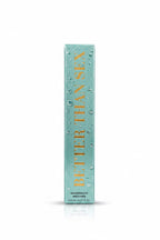 A boxed cosmetic product labeled 'Better Than Sex' stands upright on a reflective counter, with a gold promotional insert behind it. A person in a black shirt is visible in the background holding a phone, and a laptop is open on the counter.