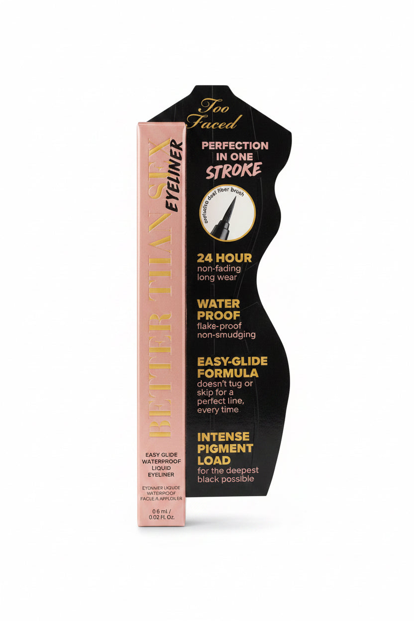 A hand holding a tube of Too Faced Better Than Sex Eyeliner against a busy desk background with a laptop and other items.