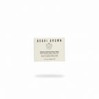 A hand holding a square cream-colored box of Bobbi Brown Vitamin Enriched Face Base, photographed in a retail or storage setting with a glass counter and other products visible in the background.