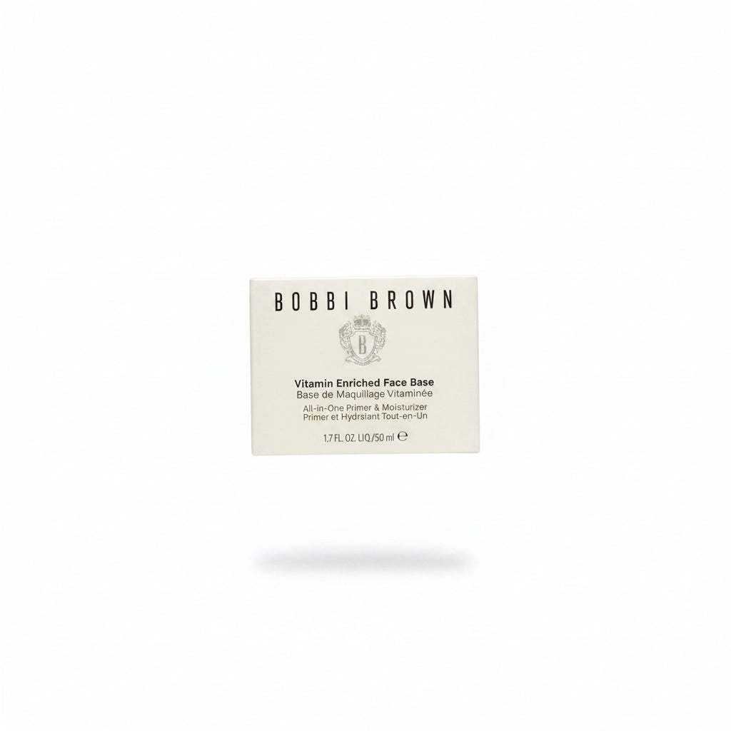 A hand holding a square cream-colored box of Bobbi Brown Vitamin Enriched Face Base, photographed in a retail or storage setting with a glass counter and other products visible in the background.