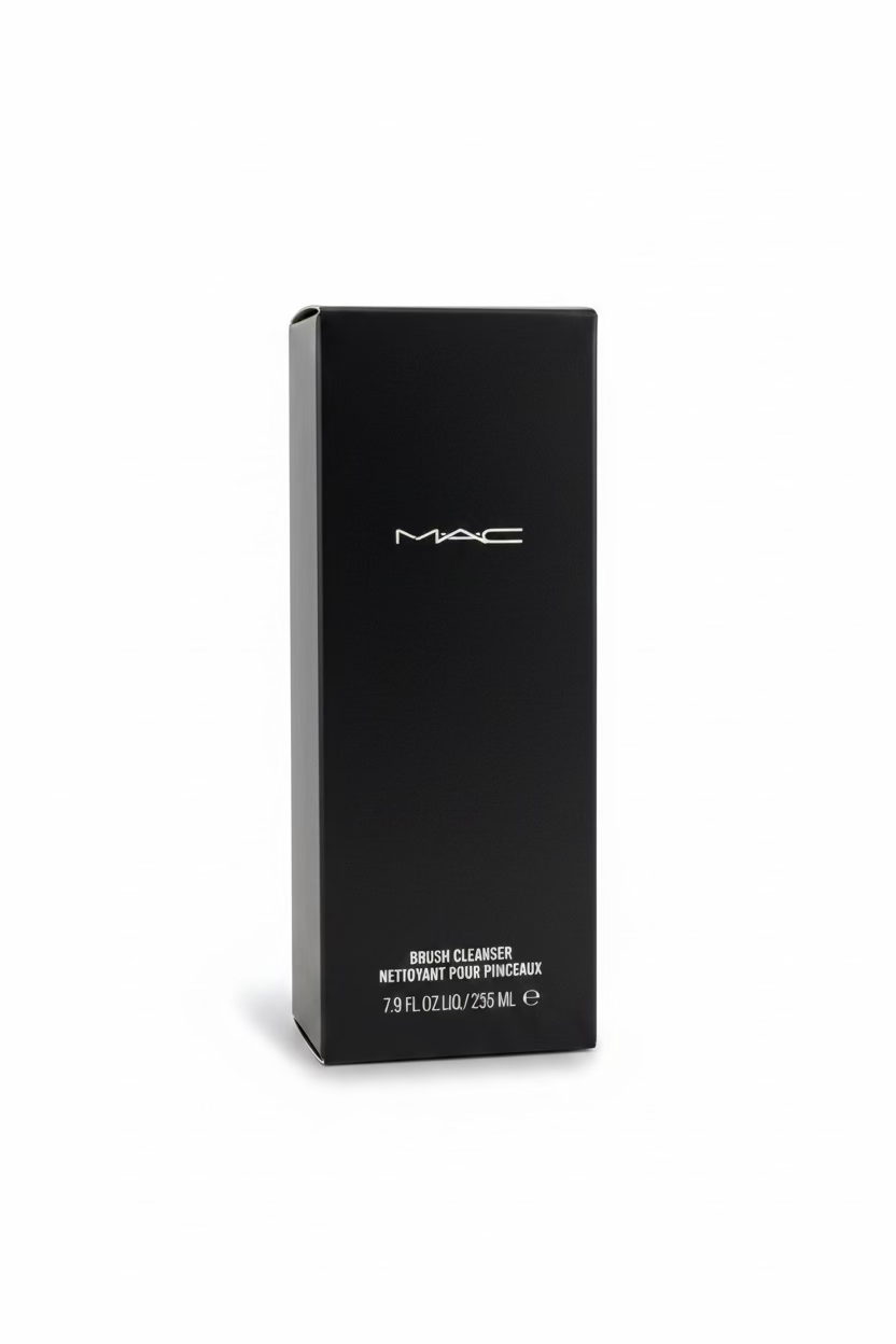 A black MAC Brush Cleanser box standing upright on a glass display counter inside a retail store. The MAC logo is printed in white on the front, with shelves of cosmetics and a laptop visible in the background.
