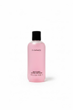 A bottle of MAC Brush Cleanser liquid makeup remover with a black cap and pale pink liquid inside, placed on a glass shelf in a retail store setting with a hand visible in the foreground.