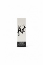 A boxed MAC Fix + Alcohol-Free Multistrating Styling Spray stands on a glass display counter in a retail cosmetics store. The tall white box has black MAC branding and large 'fix+' script.