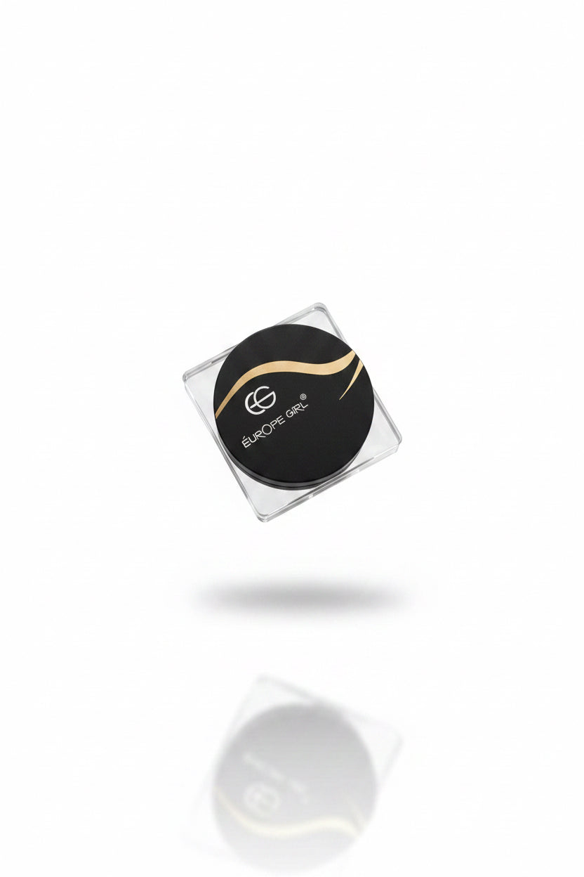 A hand holding a small square clear plastic container with a round black lid printed with the Europe One logo and text, set against a background of beige clothing and a cluttered indoor desk area.