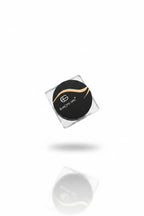 A hand holding a small square clear plastic container with a round black lid printed with the Europe One logo and text, set against a background of beige clothing and a cluttered indoor desk area.