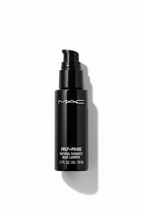 A black MAC Prep + Prime foundation bottle standing on a glass display counter in a retail setting, with a laptop and store fixtures visible in the background.
