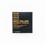 A hand holding a MAC Studio Fix concealer and corrector palette in the shade Rich Yellow Burnt Coral. The packaging is black with orange and white text, and the background shows a cluttered desk with other cosmetic items.
