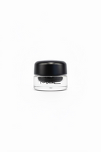 A hand holding a small clear jar of MAC Prime Color Glitter eyeshadow with a black lid, with a gold and green MAC product pouch blurred in the background.