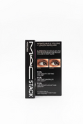 MAC Stack mascara box standing upright on a glass counter in a retail store. The black packaging shows the MAC logo, 'STACK' on the spine, and text for 'Infinite-Build Volume + Length Mascara' with before/after eye imagery and claims about the SuperStam Mega Brush.