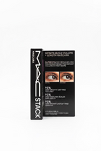 MAC Stack mascara box standing upright on a glass counter in a retail store. The black packaging shows the MAC logo, 'STACK' on the spine, and text for 'Infinite-Build Volume + Length Mascara' with before/after eye imagery and claims about the SuperStam Mega Brush.