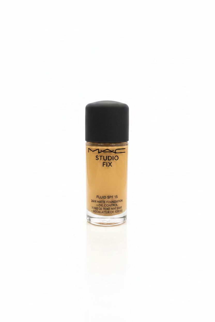 A bottle of MAC Studio Fix Fluid SPF 16 foundation with beige liquid visible through the clear bottle and a black cap, placed on a glass counter in a busy retail or studio workspace with a laptop, hand, and stacked boxes in the background.