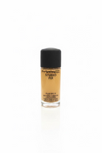 A bottle of MAC Studio Fix Fluid SPF 16 foundation with beige liquid visible through the clear bottle and a black cap, placed on a glass counter in a busy retail or studio workspace with a laptop, hand, and stacked boxes in the background.