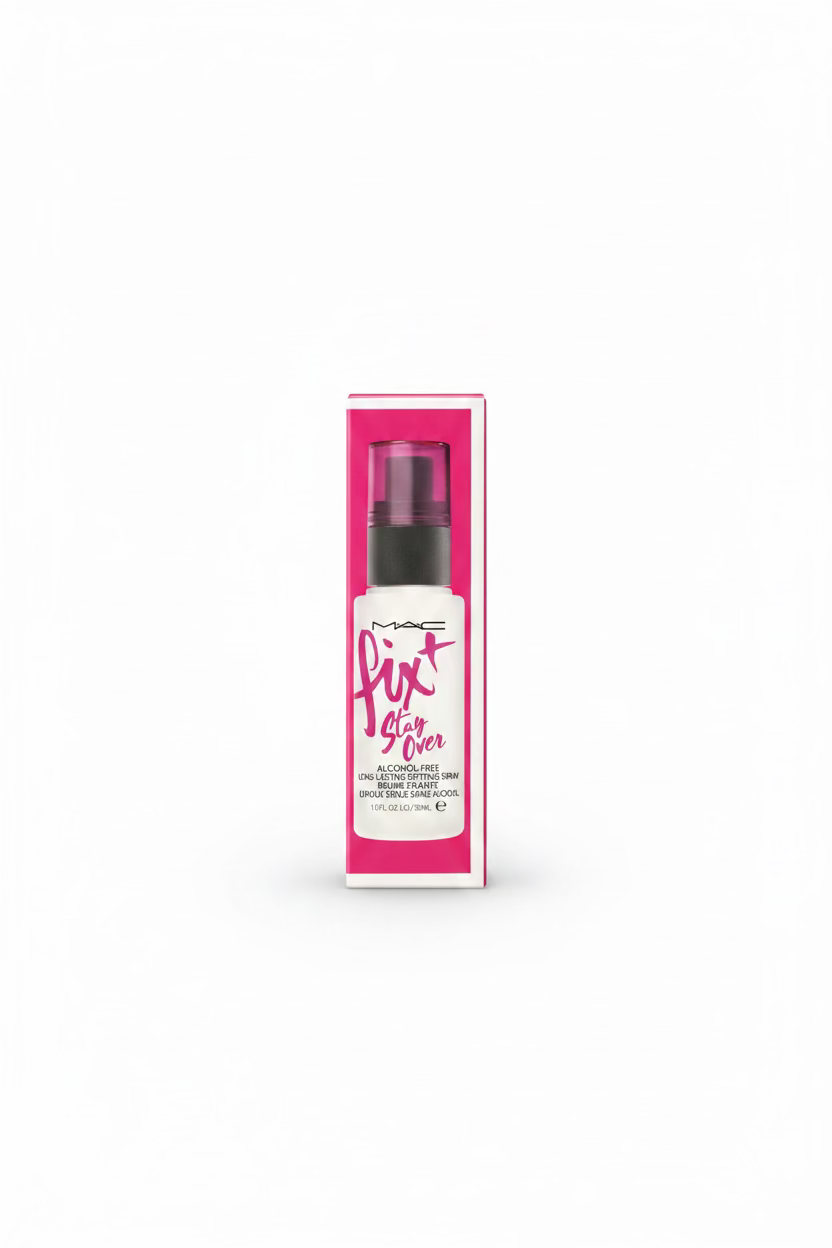A pink and white box of MAC Fix Stay Over alcohol-free oil setting spray stands on a glass display counter in a retail store. A hand is visible using a laptop in the background, with various branded boxes and store items around it.