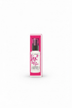 A pink and white box of MAC Fix Stay Over alcohol-free oil setting spray stands on a glass display counter in a retail store. A hand is visible using a laptop in the background, with various branded boxes and store items around it.