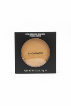 MAC Extra Dimension Skinfinish powder compact in its black retail box, displayed on a glass counter with a laptop and store shelves in the background.