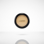 A hand holding a round black compact containing a shimmering gold eyeshadow, with a plastic-wrapped makeup item visible in the background.