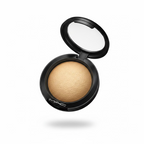 A hand holding a round black MAC eyeshadow compact showing a shimmering warm beige-gold eyeshadow powder. The background shows a cluttered storage or retail environment with jeans, plastic bags, and a desk.